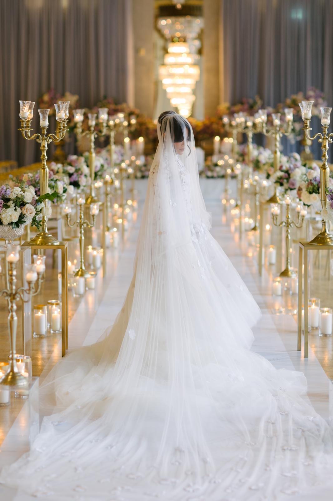 A bride standing in a candlelit ballroom aisle surrounded by lavender, blush, and ivory flowers