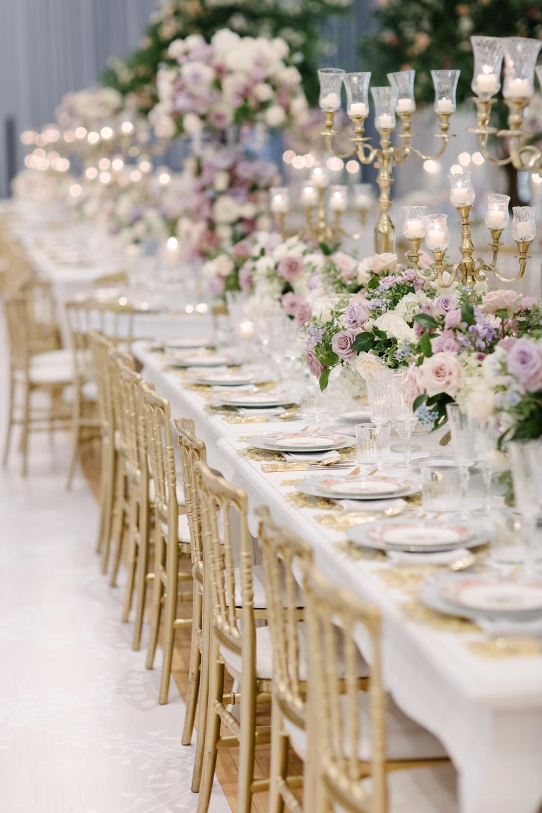 A long table layered with lavender roses, gold chairs, and candlelight
