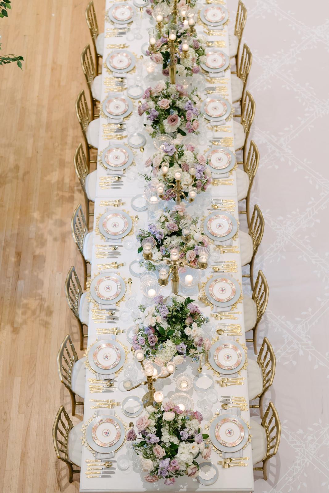 An overhead view of floral china, gold flatware, and lavender arrangements on a long table