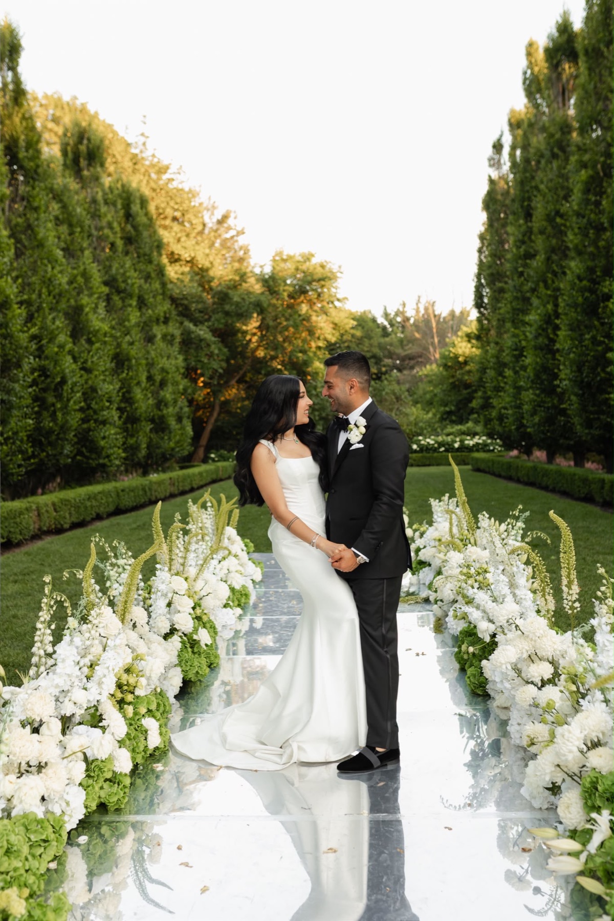 A couple together on a mirrored aisle framed by white and green flowers at Graydon Hall Manor