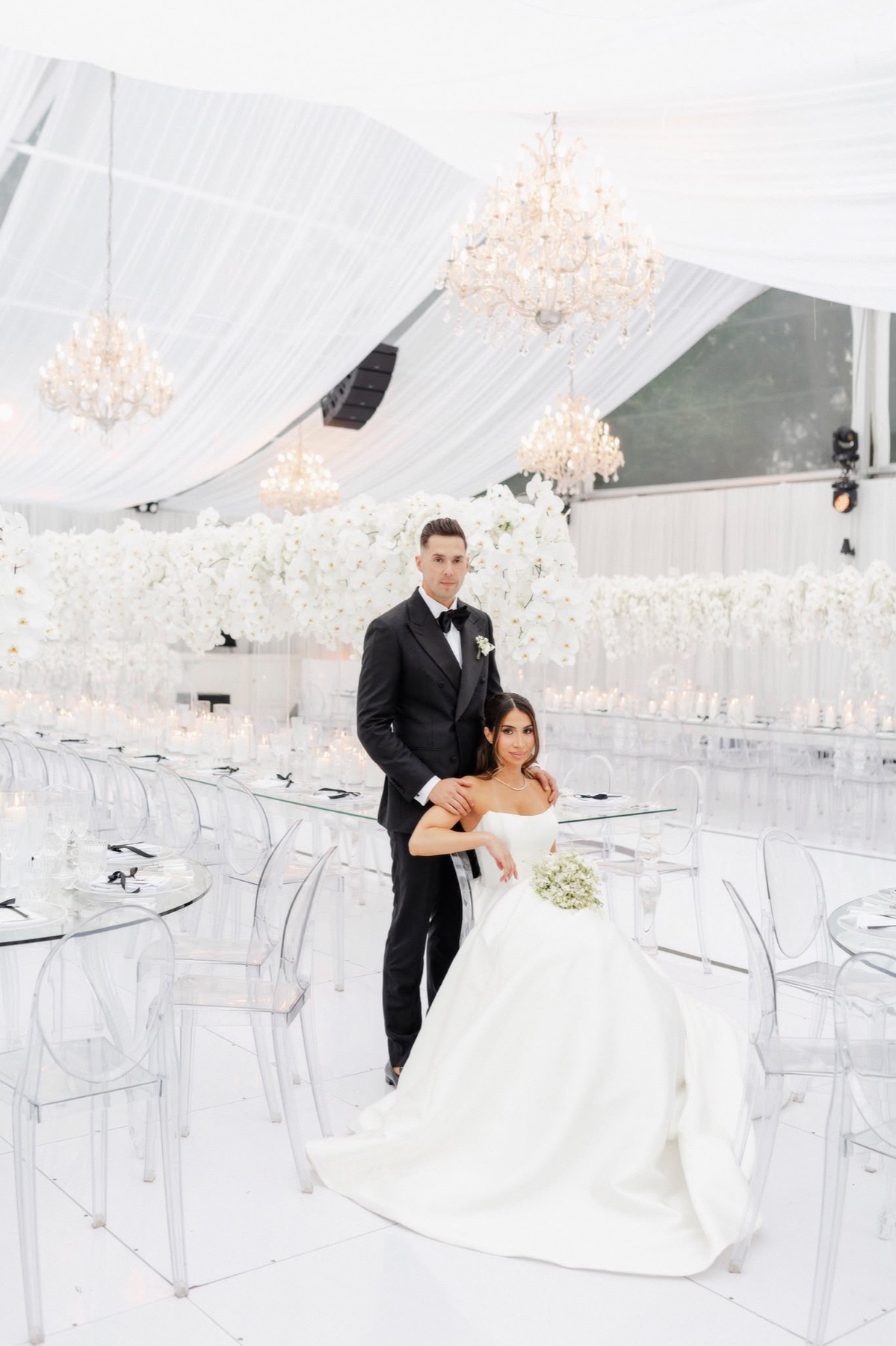 A couple posing among clear tables, white orchids, and crystal chandeliers