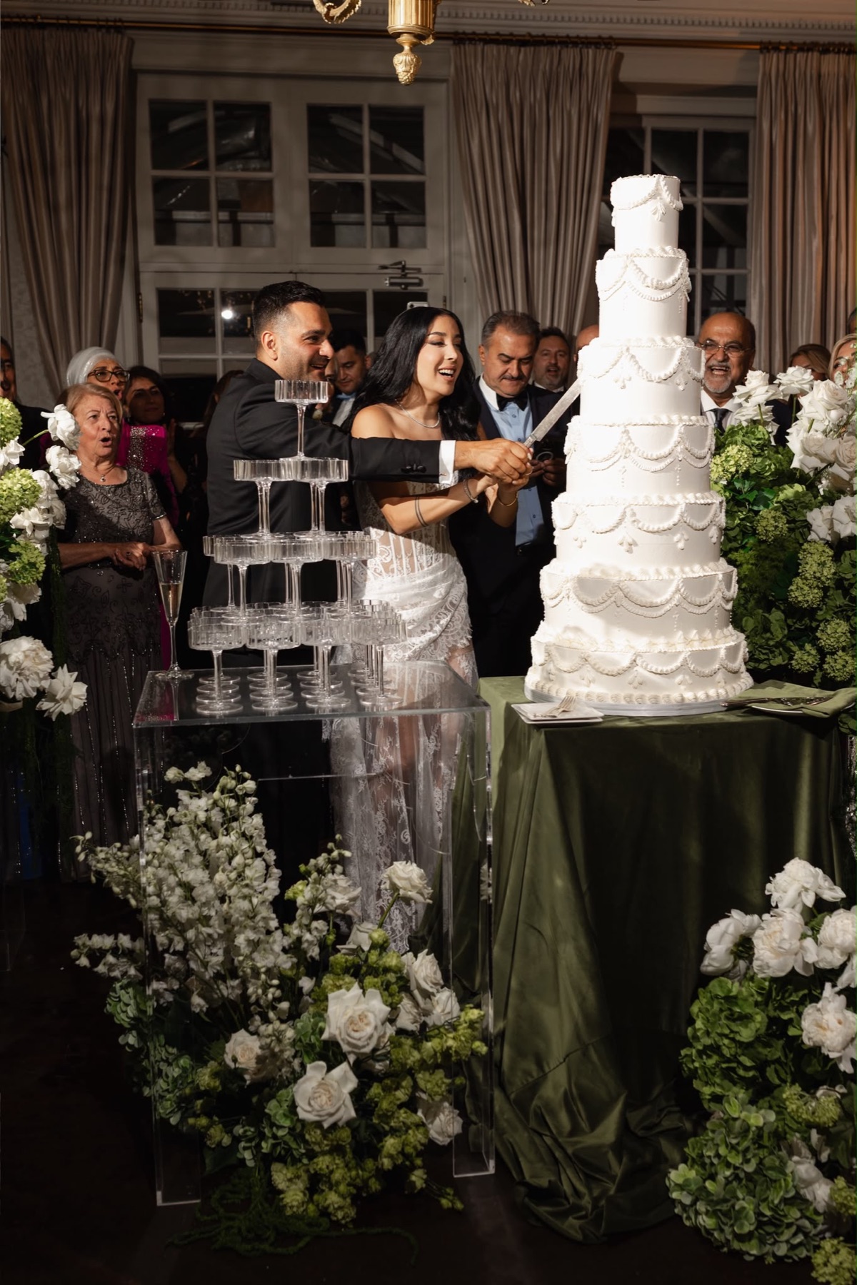 A couple cutting a tall white wedding cake surrounded by flowers