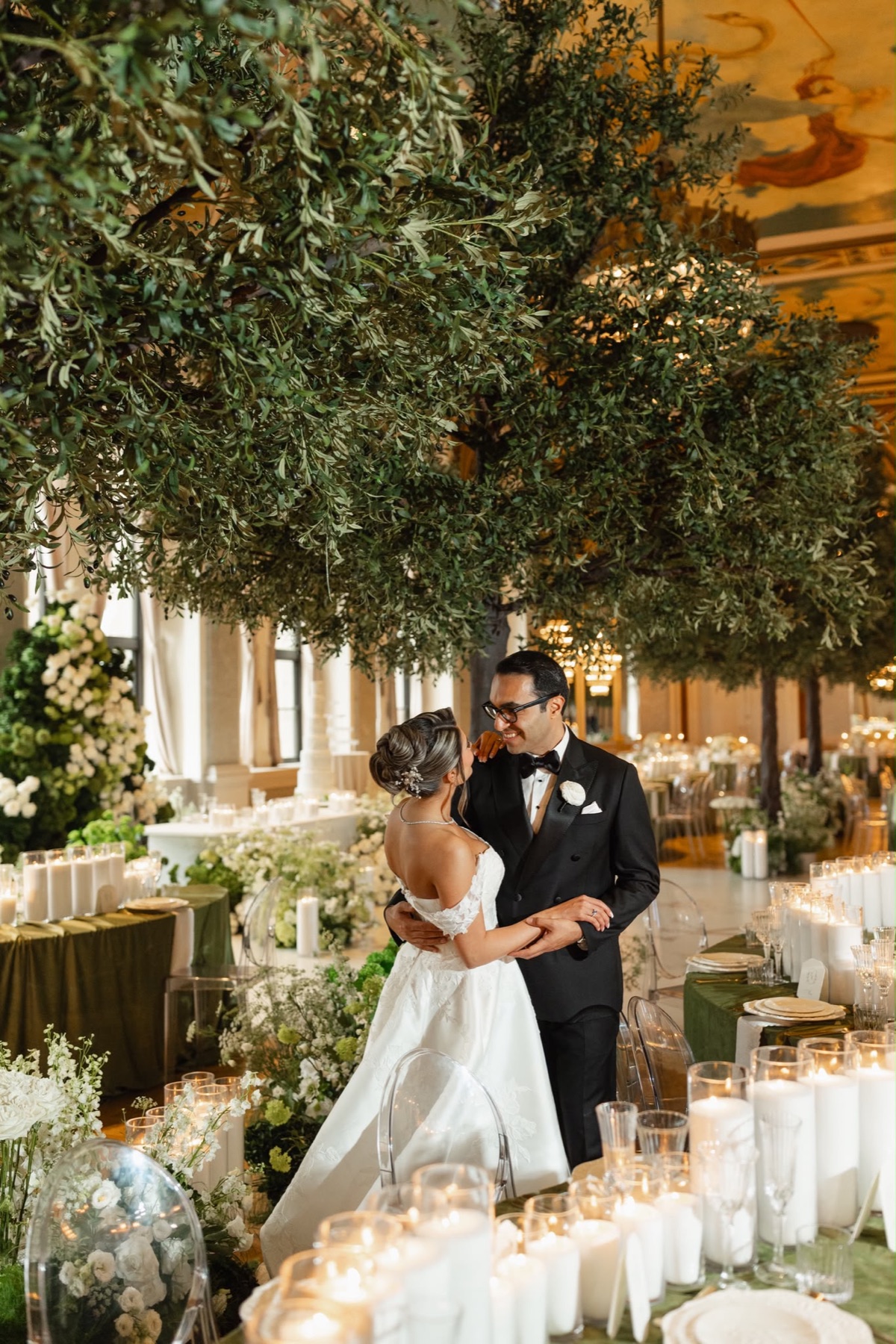 A couple dancing beneath mature indoor trees in a candlelit ballroom